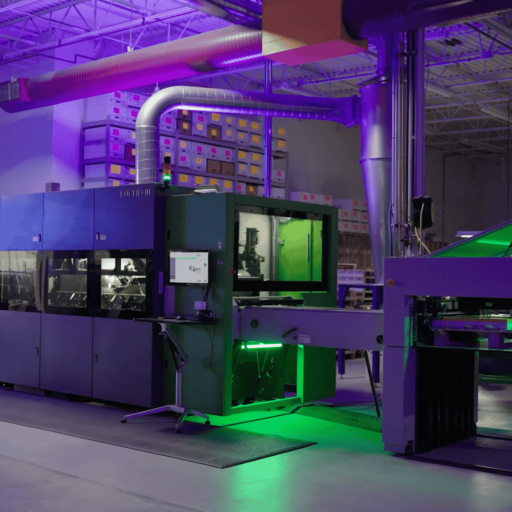 An industrial setting with complex machinery under green illumination. Ventilation pipes and shelving with boxes in the background suggest a manufacturing environment.