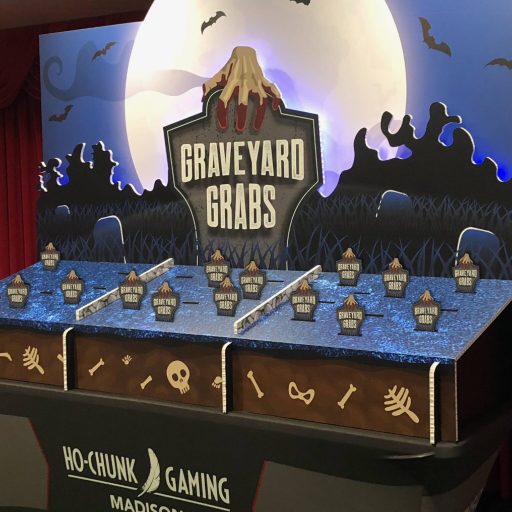 A promotional event setup labeled "Graveyard Grabs" with multiple grabber arms at a gaming venue, featuring a spooky graveyard theme with bats and a full moon.