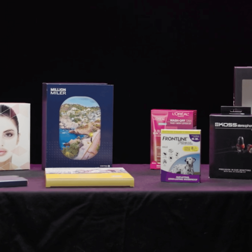 A variety of products are displayed on a table against a black background, including scents, a book, beauty items, headphones, vodka, and pet medication.
