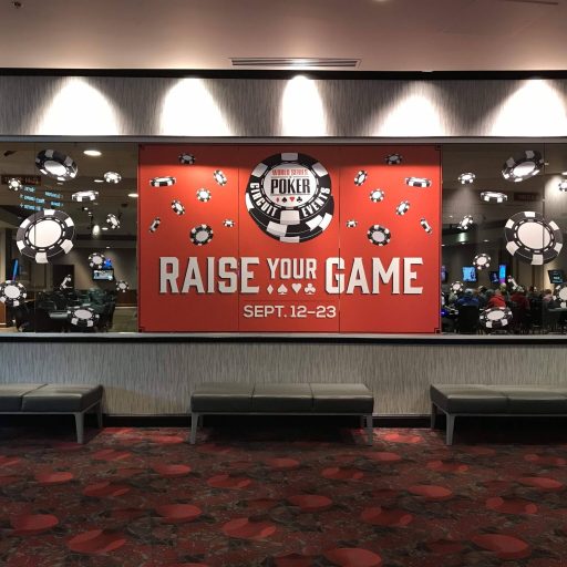 The image shows a poker room entrance with a bold "RAISE YOUR GAME" sign, people seated at tables inside, and benches in the foreground.